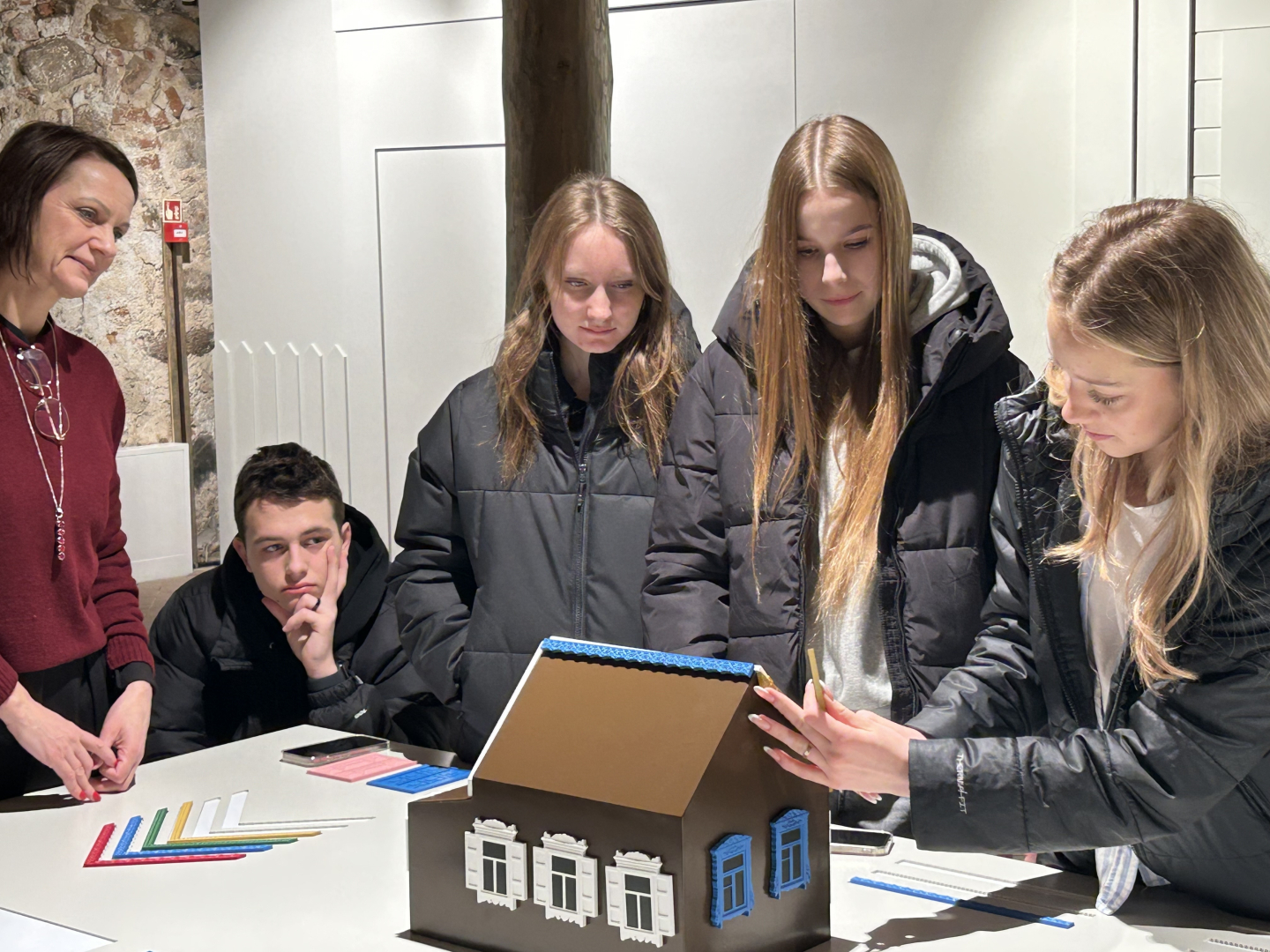 Skolēni aplūko un komplektē koka mājas modeli izstādes darbnīcā