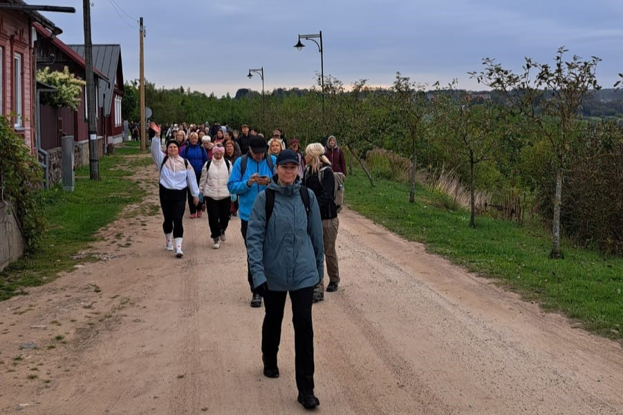 Cilvēku kolonna iet pa lauku ceļu gar mājām
