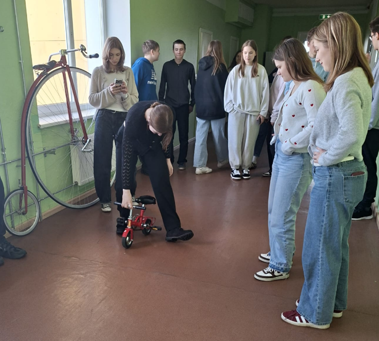 Skolēni gaitenī vēro, kā jauniete mēģina braukt ar ļoti mazu velosipēdu.