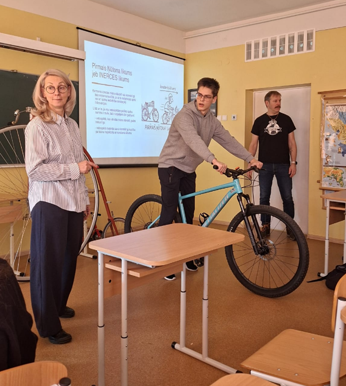 Skolēns klasē tur velosipēdu, kamēr skolotāji un citi skatās.