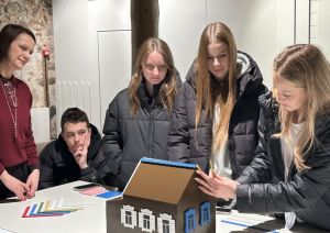 Skolēni aplūko un komplektē koka mājas modeli izstādes darbnīcā