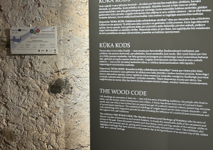 Izstādes informatīvais stends ar nosaukumu “Koka kods” uz akmens sienas fona