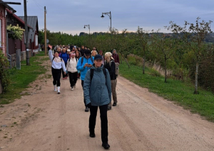 Cilvēku kolonna iet pa lauku ceļu gar mājām