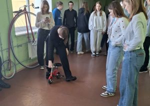 Skolēni gaitenī vēro, kā jauniete mēģina braukt ar ļoti mazu velosipēdu.