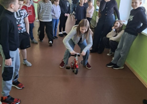 Skolēnu grupa smejas un skatās, kā meitene brauc ar mini velosipēdu gaitenī.
