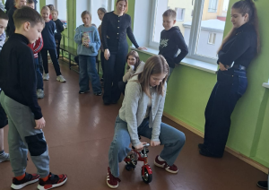 Jauniete sēž uz maza velosipēda, kamēr apkārtējie skolēni viņu vēro un smaida.