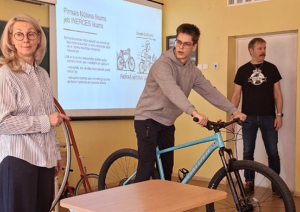 Skolēns klasē tur velosipēdu, kamēr skolotāji un citi skatās.
