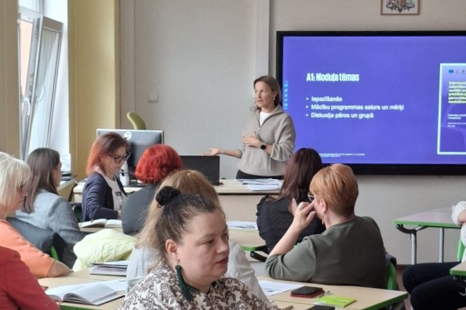 Cilvēks stāv pie ekrāna un vada nodarbību, dalībnieki sēž un klausās