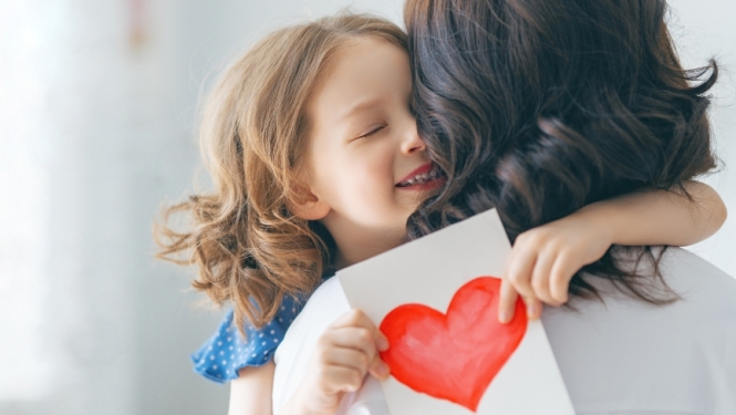 Meitene, apskāvusi mammu, tur rokās kartiņu ar uzzīmētu sirdi