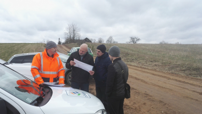4 cilvēki pēta ceļa projektu, stāvot pie mašīnas uz lauku ceļa
