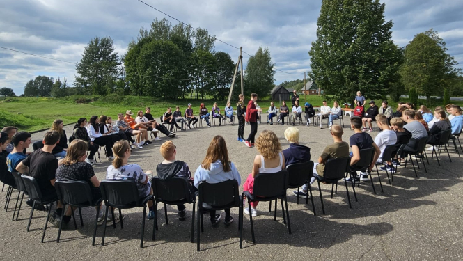 Jaunieši sasēdušies aplī uz krēsliem, ārā, siltajā laikā.