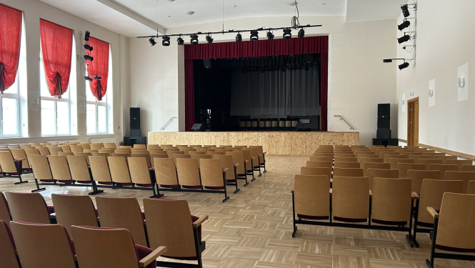 Zāle ar krēsliem, skats uz skatuvi ar aizkariem.