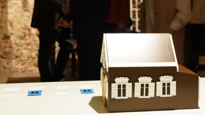 Koka mājas modelis ar dekoratīviem logiem uz galda, fonā izstādes apmeklētāji