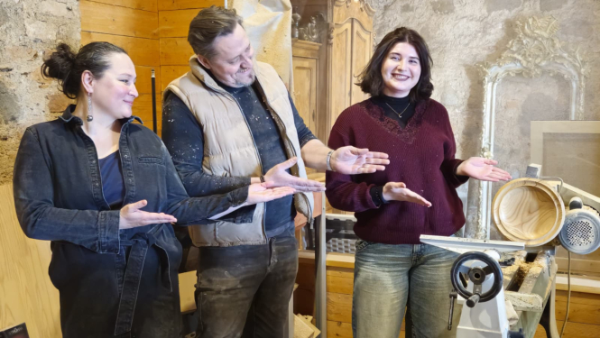 Trīs cilvēki darbnīcā prezentē koka virpošanas darbu