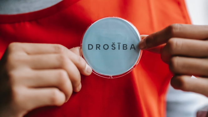 Sievietes rokas tur pie krūtīm piespraustu piespraudi ar uzrakstu "DROŠĪBA"