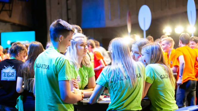 Daudz jauniešu, sadalīti grupiņās, stāv pie apaļiem galdiem