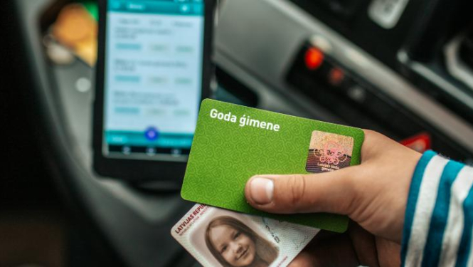 Bērns tur rokās savu "Goda ģimenes" apliecību un eID karti pie sabiedriskā transporta norēķinu termināļa.