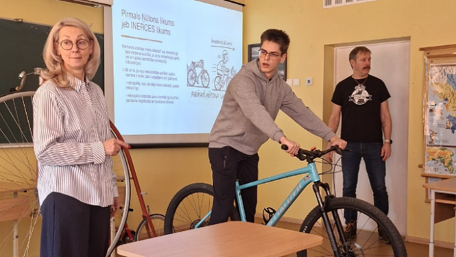 Skolēns klasē tur velosipēdu, kamēr skolotāji un citi skatās.