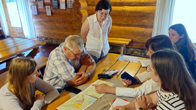 Cilvēki sēž pie galda, uz kura noliktas kartes ar teritorijas plānojumiem, kurus cilvēki apspriež