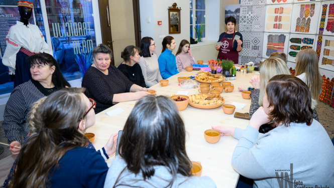 Cilvēki sēž pie galda, uz kura ir tēja un uzkodas, un klausās stāstījumu Vienības nama Tradīciju mājā