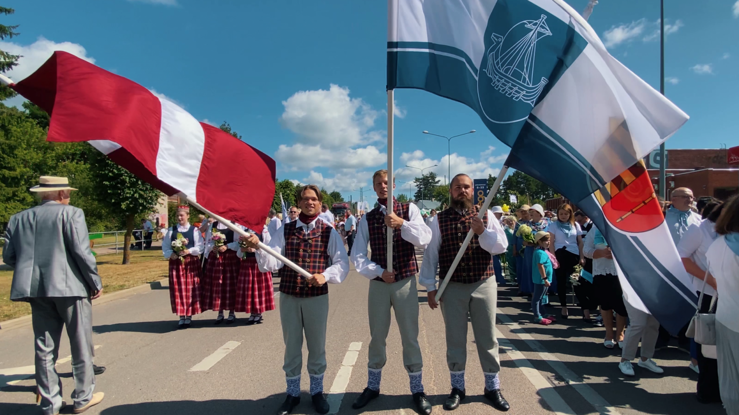 Trīs puiši tautastērpos tur Latvijas, Krāslavas un Krāslavas novada karogus. Fonā cilvēku pulks