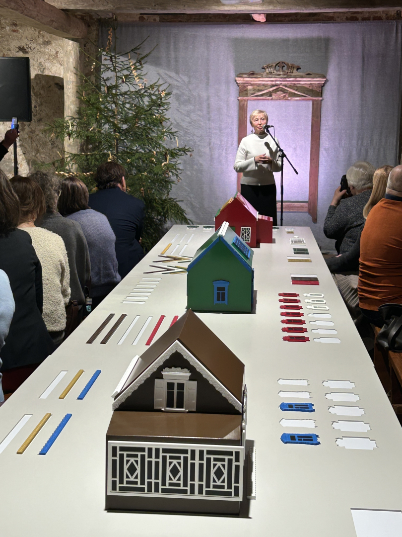 Sieviete runā pie mikrofona, priekšplānā galdi ar māju modeļiem un dekoratīviem elementiem