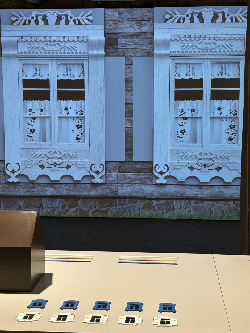 Projekcija ar tradicionāliem koka logu rotājumiem un priekšā izlikti logu detaļu paraugi