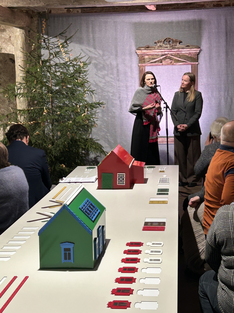 Divas sievietes uzstājas izstādes atklāšanā pie mikrofona, priekšplānā māju modeļi