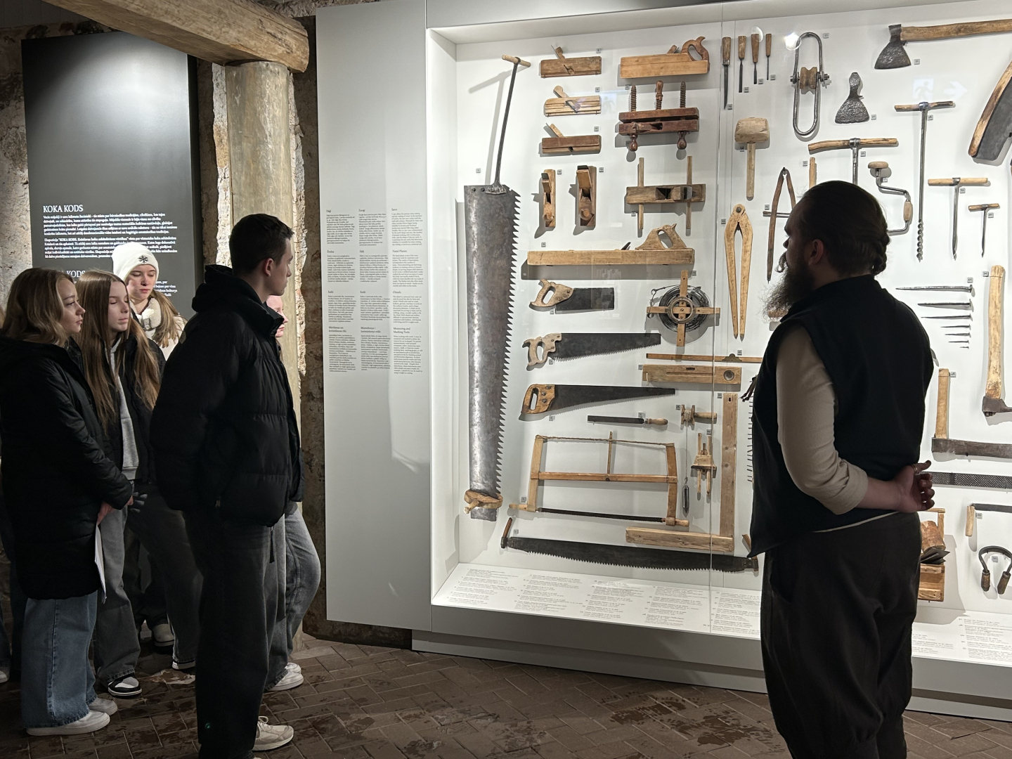 Skolēnu grupa gida pavadībā aplūko vēsturiskus kokapstrādes instrumentus vitrīnā