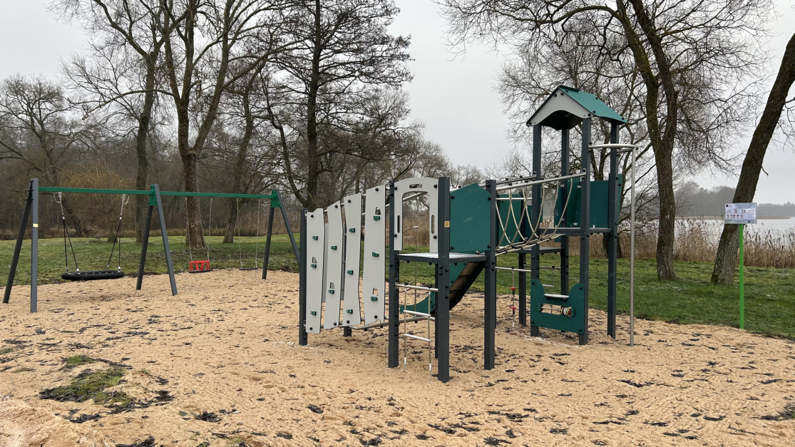 Bērnu rotaļu laukums pie ezera – kāpelēšanas konstrukcija un šūpoles smilšu laukumā