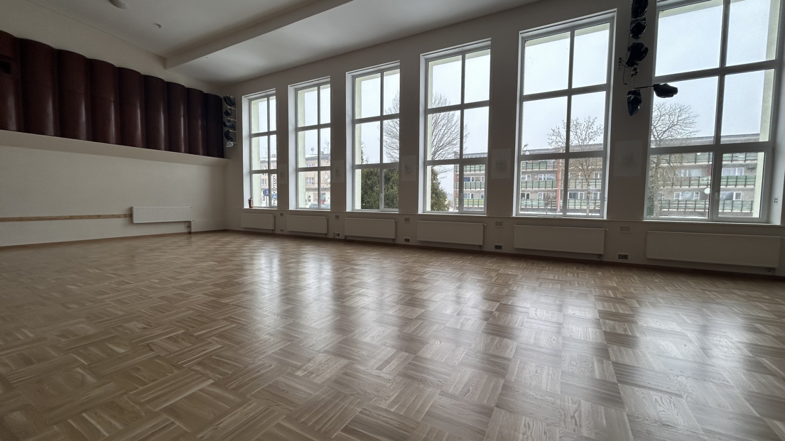 Plaša tukša zāle ar parketa grīdu un lieliem logiem.