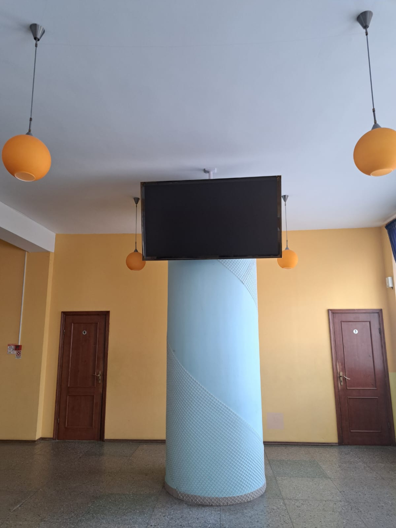 Kolonna ar piekārtu ekrānu un durvīm fonā.