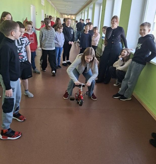 Skolēnu grupa smejas un skatās, kā meitene brauc ar mini velosipēdu gaitenī.