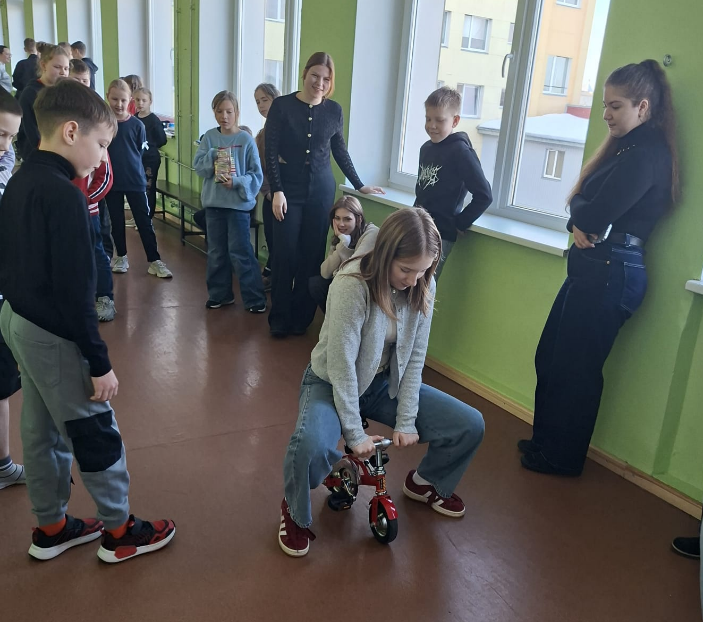 Jauniete sēž uz maza velosipēda, kamēr apkārtējie skolēni viņu vēro un smaida.