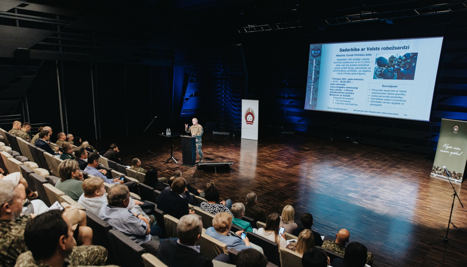 Cilvēki sēž zālē un klausās Valsts robežsardzes prezentāciju
