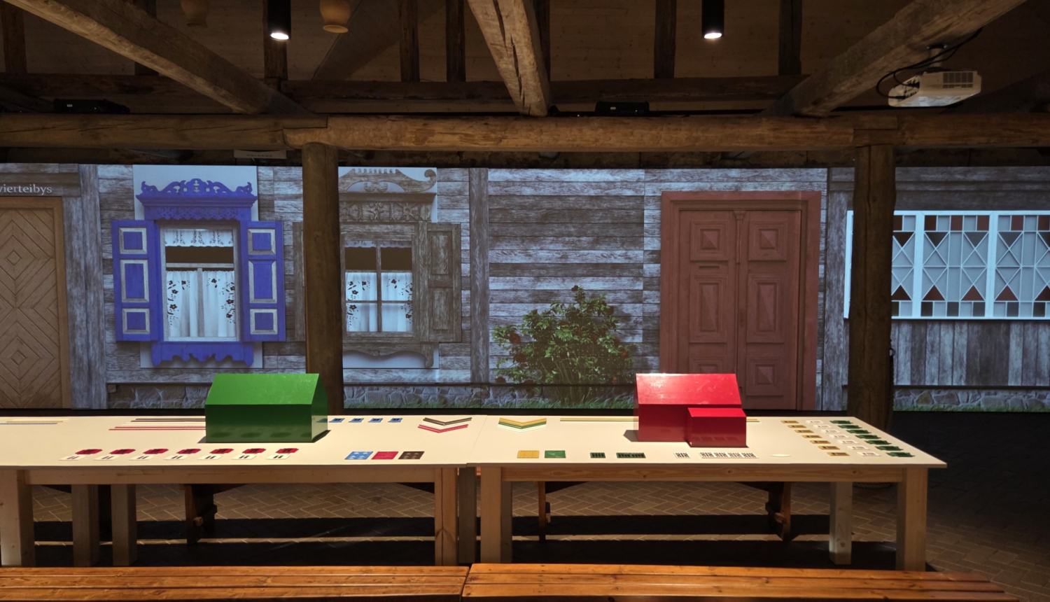 Interaktīva izstāde, uz galdiem novietoti krāsaini maketi un kartītes, fonā projicēta tradicionālas koka mājas fasāde