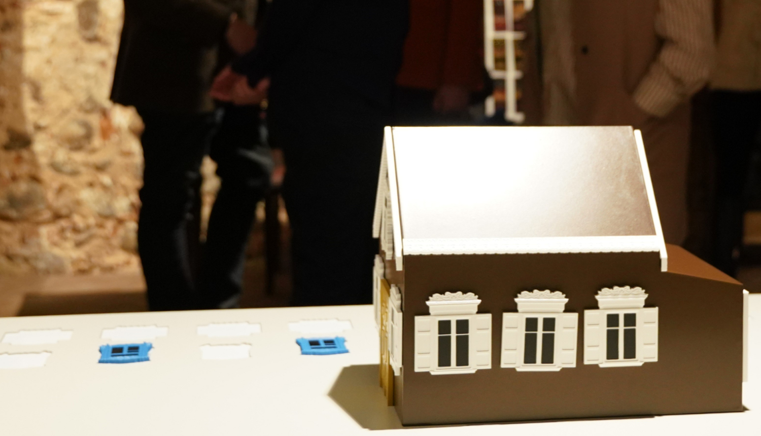 Koka mājas modelis ar dekoratīviem logiem uz galda, fonā izstādes apmeklētāji