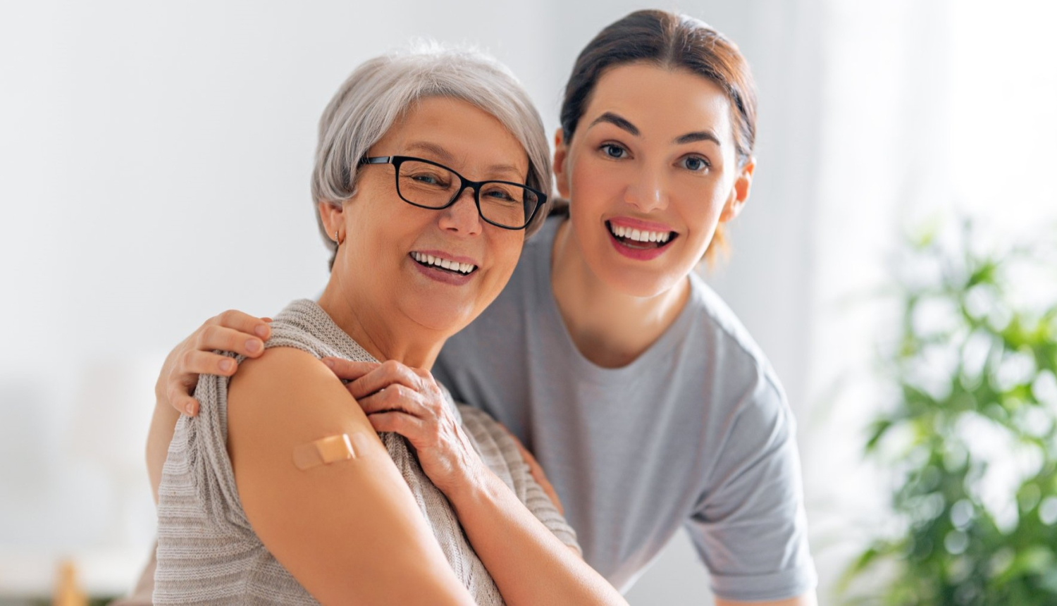 Jauna, smaidīga sieviete apskauj priecīgu seniori pēc vakcinācijas