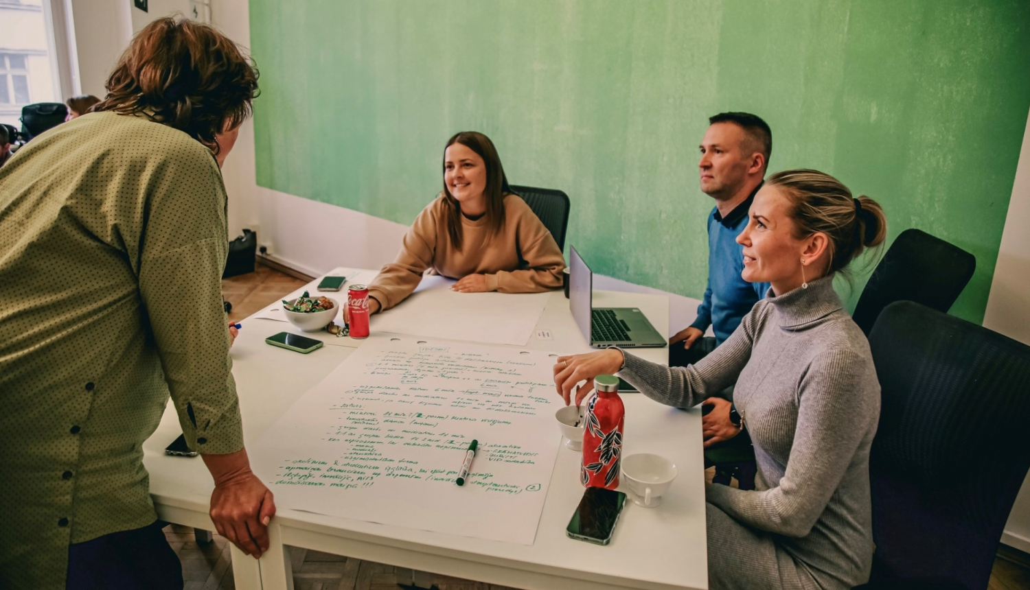 Cilvēki sēž un stāv apkārt galdam, apspriež idejas