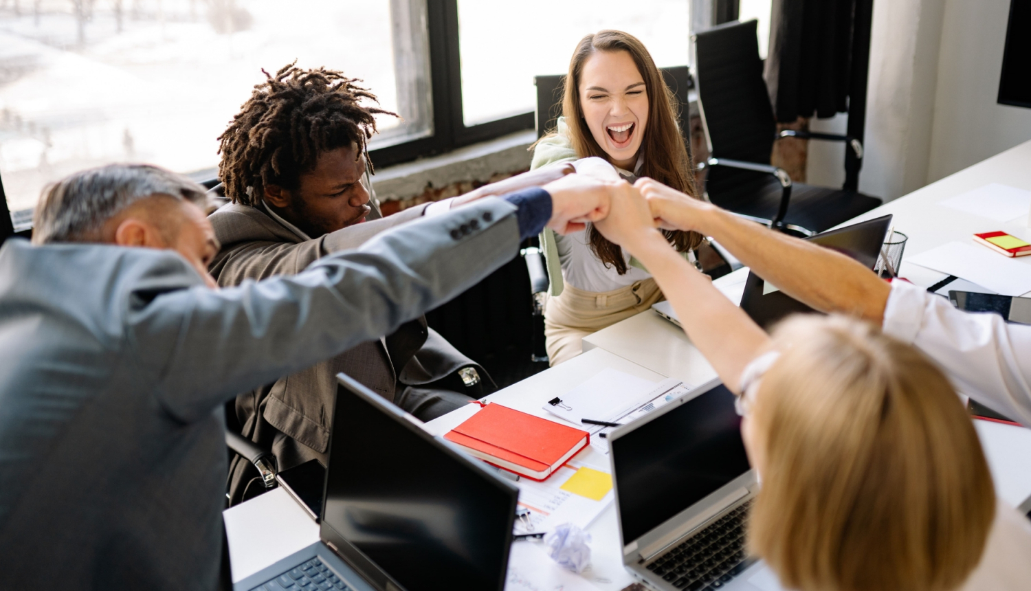 Cilvēki, sēžot pie galda, izstiepj savas rokas lai pieskartos viens otram ar dūrēm