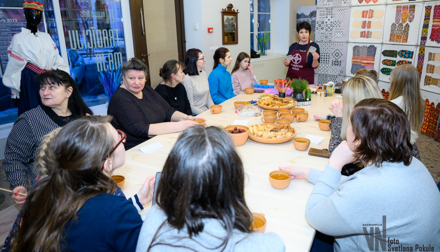 Cilvēki sēž pie galda, uz kura ir tēja un uzkodas, un klausās stāstījumu Vienības nama Tradīciju mājā