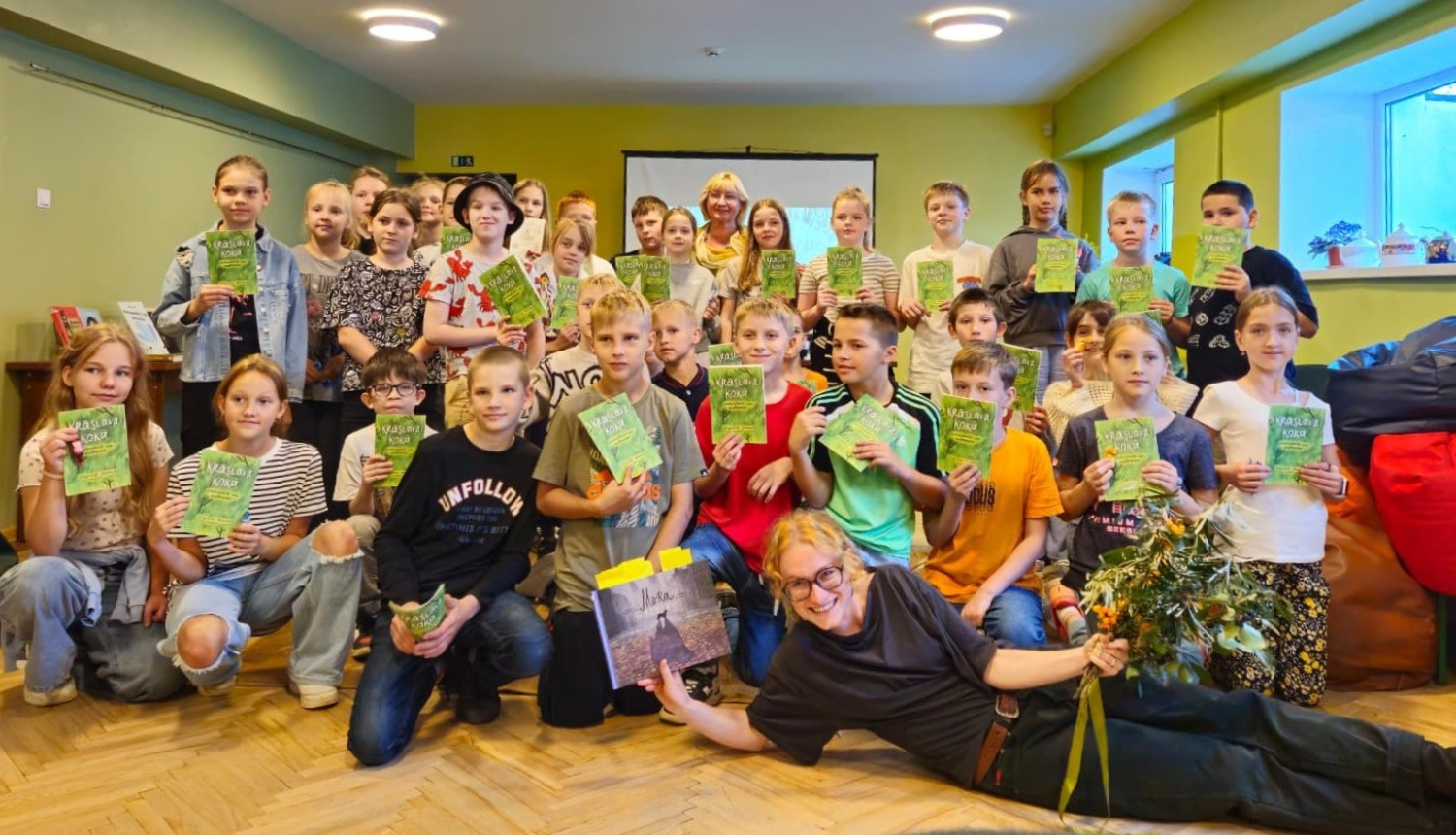 Liela bērnu grupa ar ilustratori Martu Folkmani Krāslavas novada bibliotēkā, katram rokās grāmata