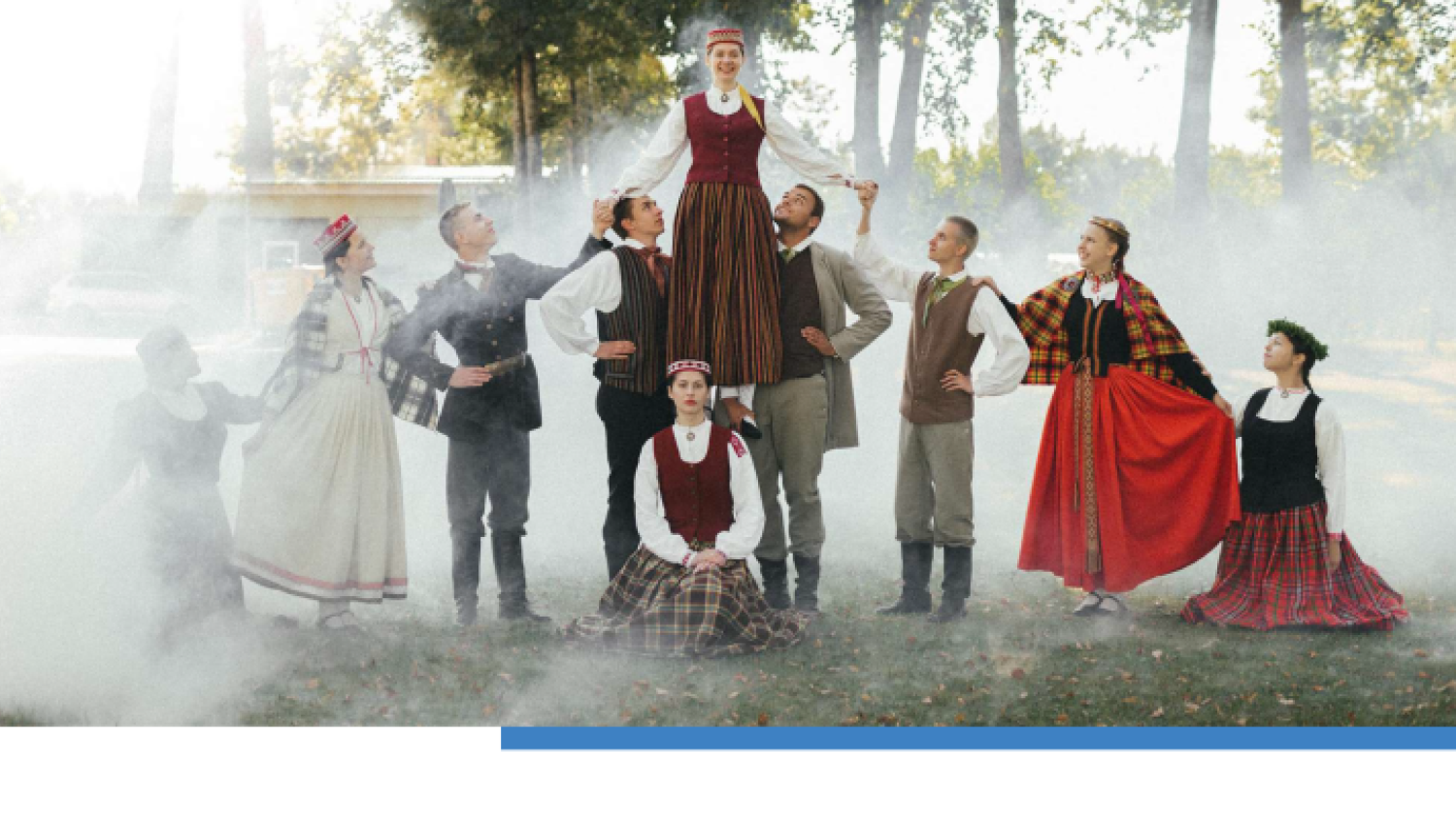 Cilvēku grupa latviešu tautastērpos stāv meža ielokā miglā. Projekta publicitātes plakāts.