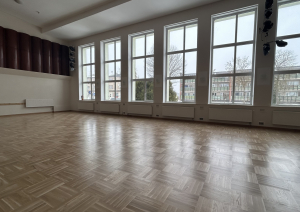 Plaša tukša zāle ar parketa grīdu un lieliem logiem.