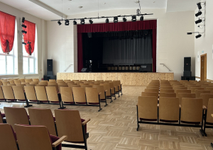 Zāle ar krēsliem, skats uz skatuvi ar aizkariem.